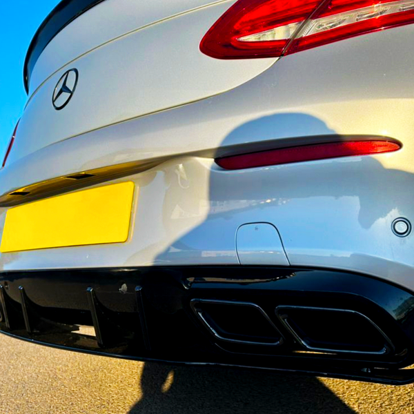 A silver car's rear bumper with a yellow license plate and dual exhaust is parked on a road. The context includes a clear blue sky and sunlight casting shadows.