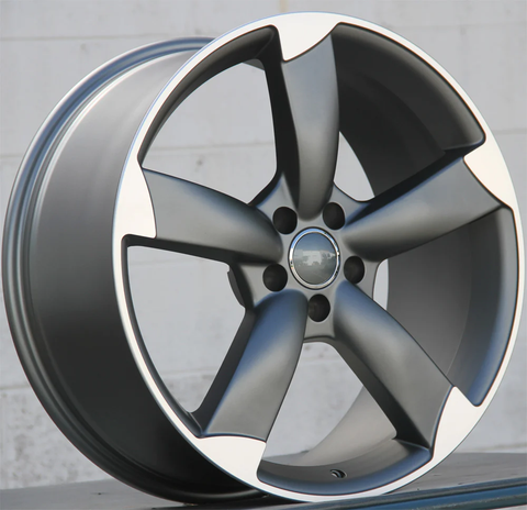 A metallic car wheel rim with five wide spokes is positioned upright. It's placed on a reflective surface in front of a neutral, light-colored wall.