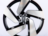 A five-spoke alloy wheel, featuring a polished black and silver design with a central logo, is positioned upright on a plain, light background.