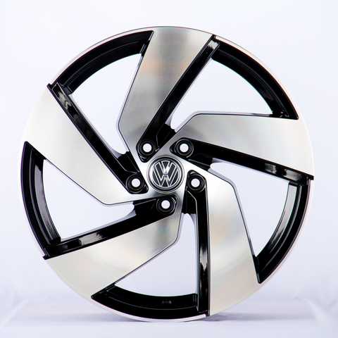 A five-spoke alloy wheel, featuring a polished black and silver design with a central logo, is positioned upright on a plain, light background.