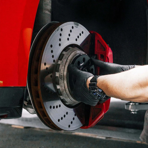 A gloved hand is installing a ventilated brake disc on a car's wheel hub. The car is elevated, showing a red brake caliper in a garage setting.