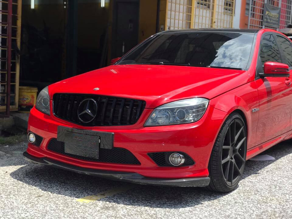 Red Mercedes-Benz car parked on a sunlit street, featuring black rims and tinted windows. Nearby, there are buildings with yellow grids and a partially open garage door.