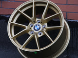 A gold BMW alloy wheel rests against a brick wall on a rough black surface. Center logo reads 