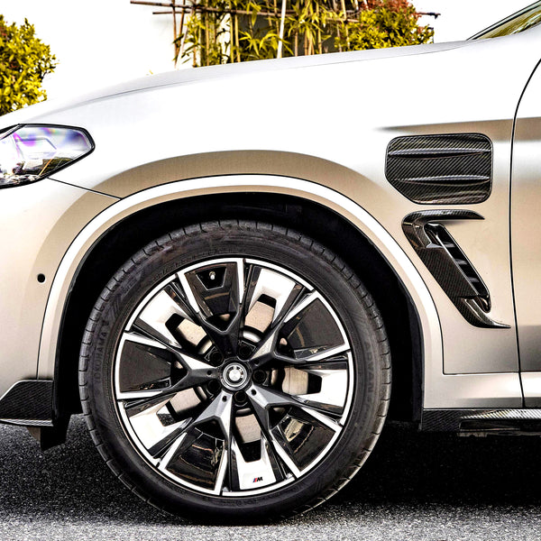 A silver car's front tire, featuring intricate alloy wheel design, is parked on a street. Greenery lines the background, providing a natural contrast to the vehicle.