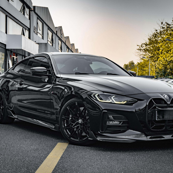 A sleek black sports car parked on a street, with a reflective body and distinctive headlights, surrounded by modern buildings and trees under a clear sky.