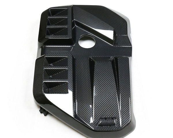 A carbon fiber engine cover with geometric patterns and a circular cutout, placed on a plain white background.