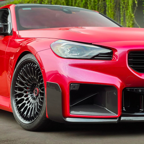 A sleek, red sports car with intricate black rims is parked on a smooth surface, surrounded by lush greenery. The front headlight and grill are prominently featured.