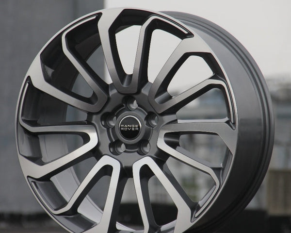 A modern alloy wheel stands upright, displaying intricate split-spoke design. Center hub bearing the text "RANGE ROVER." Positioned in an outdoor, urban setting with a concrete base.