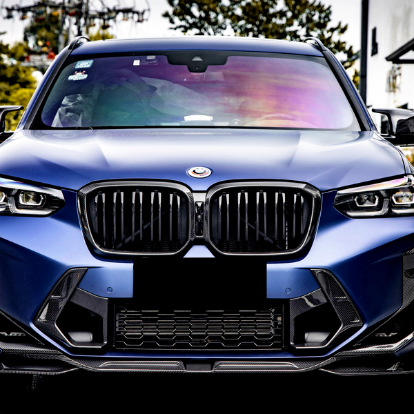 A blue SUV parked on a street displays a glossy front grille and headlights. Surrounding the car are trees, parked cars, and a building, creating an urban environment.