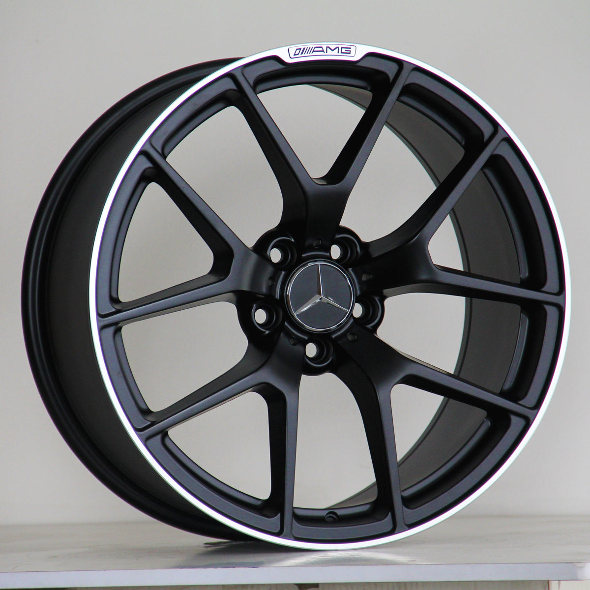 A black alloy wheel with a five-spoke design, featuring a Mercedes-Benz logo in the center and "AMG" text on the rim, rests against a simple, plain background.