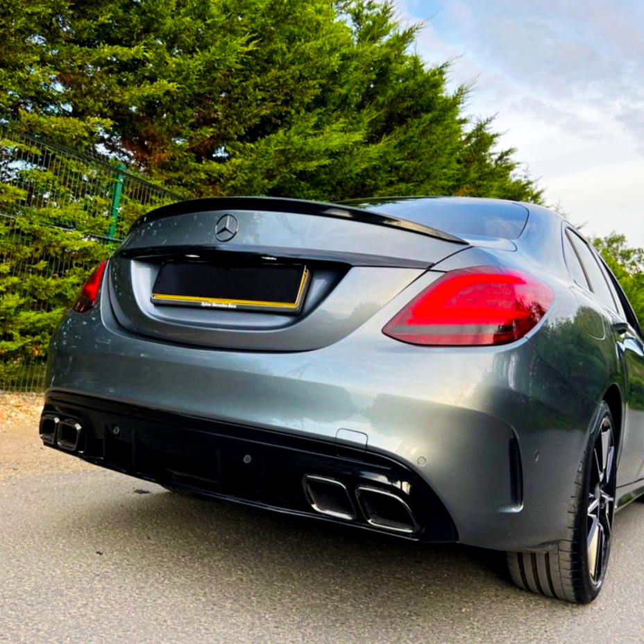 A gray Mercedes-Benz car is parked on a road, surrounded by green trees and a wire fence in the background. The car's rear license plate is blacked out.