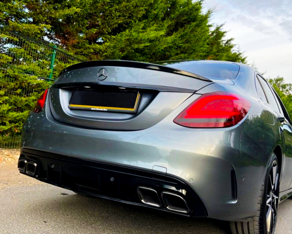 A gray Mercedes-Benz car is parked on a road, surrounded by green trees and a wire fence in the background. The car's rear license plate is blacked out.