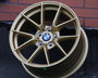 A gold BMW wheel rim leans against a brick wall on a dark pavement. The BMW logo is visible at the center. Text on the rim reads: "flow forming process."