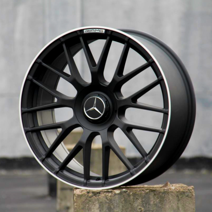 A black alloy wheel with Mercedes-Benz logo and "AMG" text is displayed upright on concrete blocks against a blurred, neutral background.