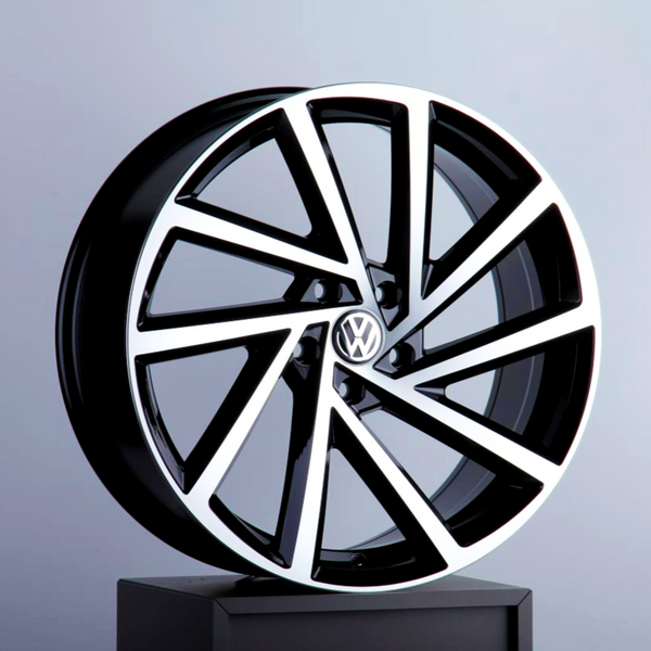 A polished, black-and-silver car wheel rim is displayed upright, featuring a VW logo at its center, against a softly lit, plain gray background.