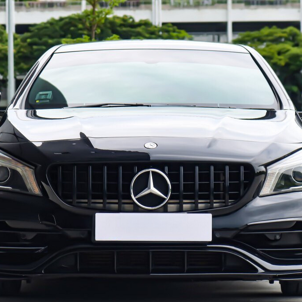 A black Mercedes-Benz car is parked outdoors, facing forward. The background consists of greenery and a multi-story building.