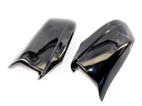 Two glossy black plastic car mirror covers, positioned side by side, reflecting light on a plain white background.