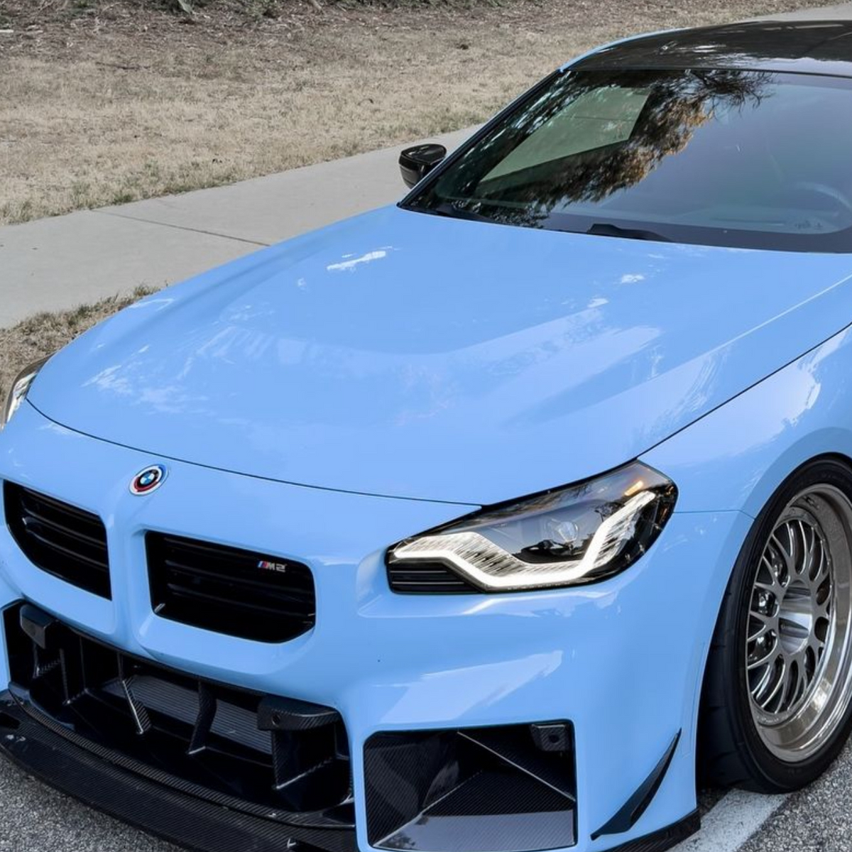 A sleek, blue sports car is parked on a road next to a grassy area, featuring a glossy black roof and distinctive front bumper with an "M2" badge.