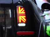 Rear taillight with a Union Jack pattern glows brightly on a black car. Reflections and surrounding vehicles indicate an indoor or dimly lit environment, possibly a parking garage.