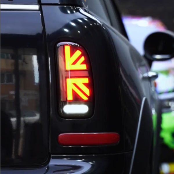Rear taillight with a Union Jack pattern glows brightly on a black car. Reflections and surrounding vehicles indicate an indoor or dimly lit environment, possibly a parking garage.