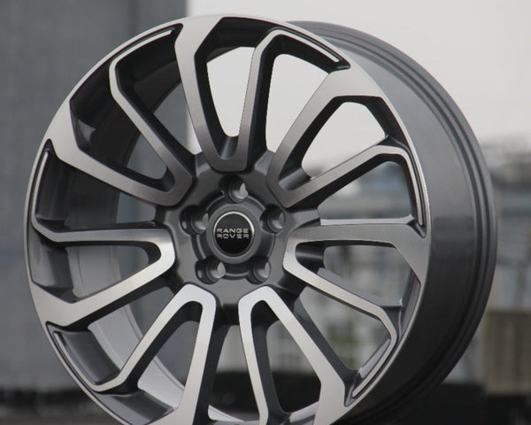 A polished silver and black alloy wheel rests upright on concrete. "Range Rover" is inscribed at its center. The background is blurred, suggesting an urban setting.