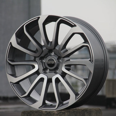 A polished silver and black alloy wheel rests upright on concrete. "Range Rover" is inscribed at its center. The background is blurred, suggesting an urban setting.