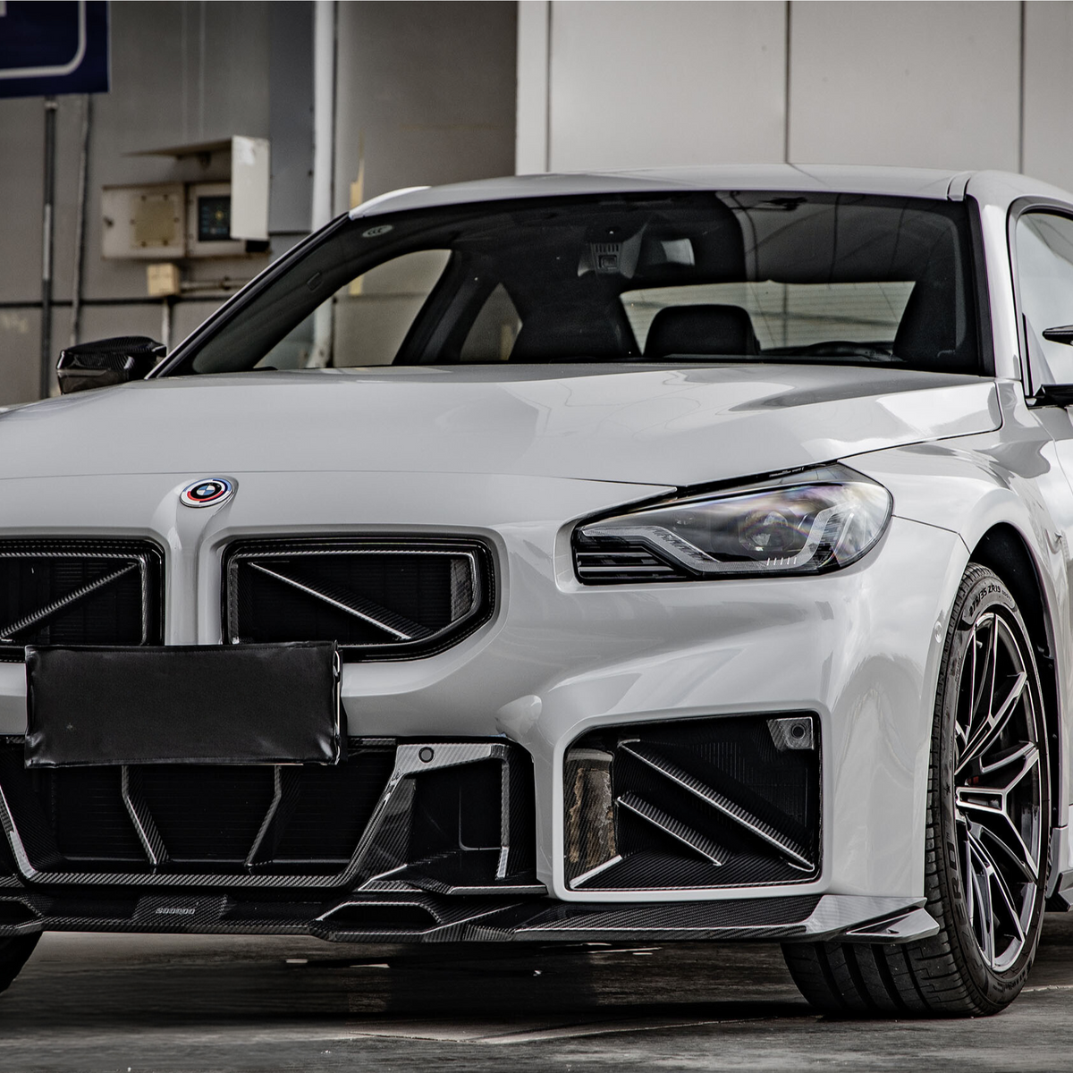 A white sports car is parked in a garage, displaying a sleek body with aggressive front styling. The setting includes a wall-mounted parking sign and exposed industrial elements.