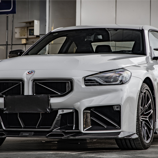 A white sports car is parked in a garage, displaying a sleek body with aggressive front styling. The setting includes a wall-mounted parking sign and exposed industrial elements.