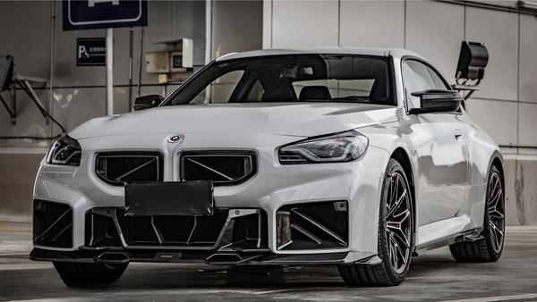 A white sports car is parked in a garage, displaying a sleek body with aggressive front styling. The setting includes a wall-mounted parking sign and exposed industrial elements.