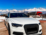 A white Audi car is parked on a tiled surface, with snow-covered mountains and a red truck in the background. The word 