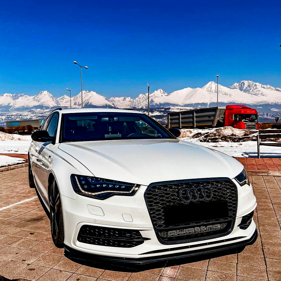 A white Audi car is parked on a tiled surface, with snow-covered mountains and a red truck in the background. The word "quattro" is visible on the car's grille.