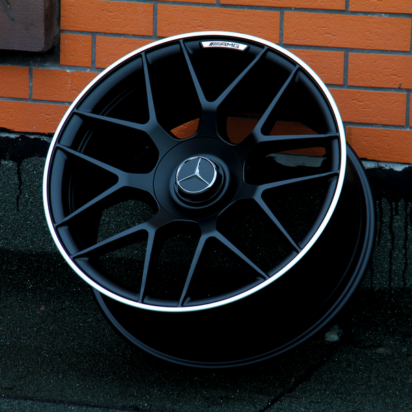 A black alloy wheel with a Mercedes-Benz logo and "AMG" text rests against an orange brick wall on a concrete surface.