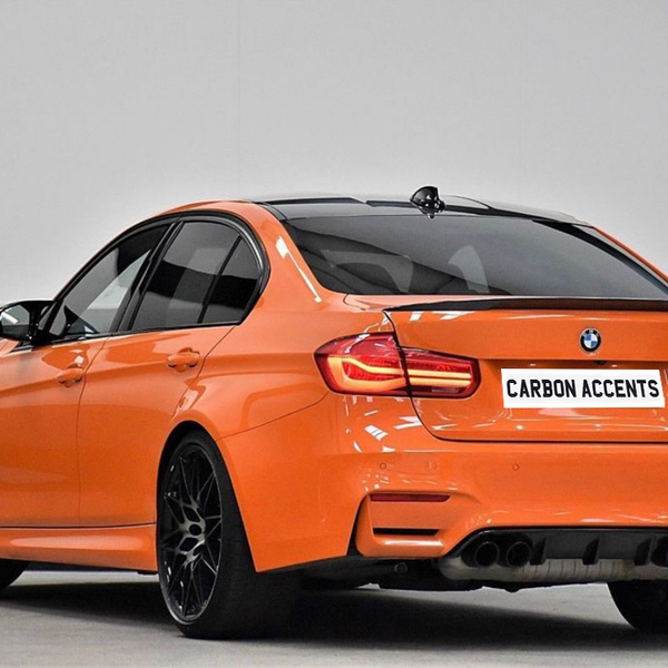 An orange BMW M3 is parked indoors, showcasing a sleek design and black wheels. The license plate reads "CARBON ACCENTS," emphasizing its sporty appeal in a neutral setting.