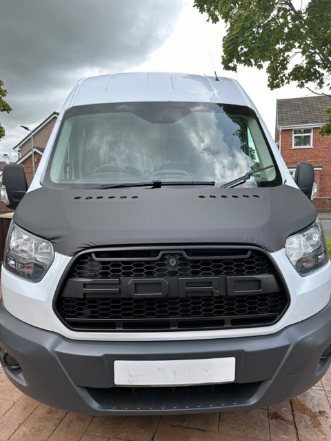 A white Ford van is parked on a brick driveway. The hood is covered with a black protective wrap, with "FORD" displayed prominently on the grille. Houses and trees surround the scene.
