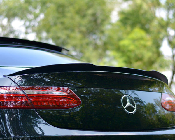 A black Mercedes-Benz car is parked outdoors, its rear showcasing a sleek spoiler and distinctive taillights. The green background of trees suggests a serene, natural setting.