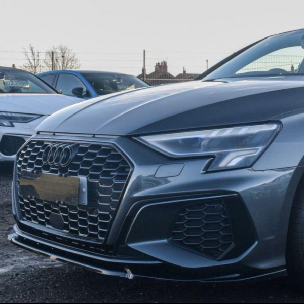 A grey Audi car is parked on a gravel lot. Its front grille is prominent, adjacent to another white car. A fence and trees are visible in the background.