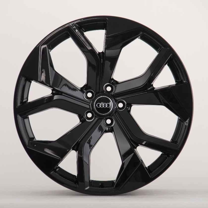 A glossy black car wheel features a five-spoke design with intersecting shapes, prominently displaying the Audi logo at the center on a plain, light background.