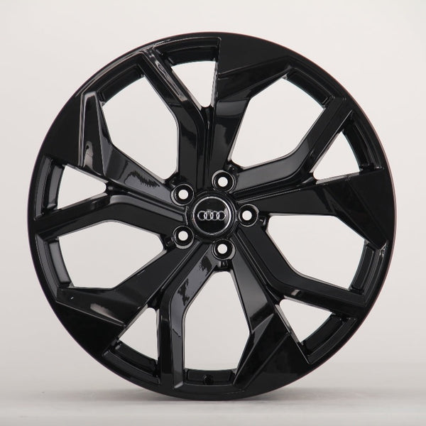 A glossy black car wheel features a five-spoke design with intersecting shapes, prominently displaying the Audi logo at the center on a plain, light background.