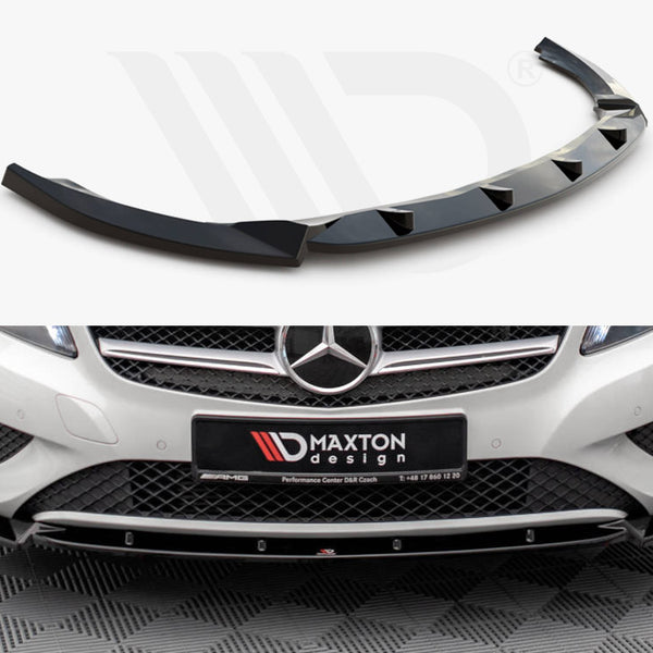 A glossy, black car splitter with angular fins is displayed alone above the grille and bumper of a white Mercedes-Benz in a showroom. License plate reads "MAXTON design."