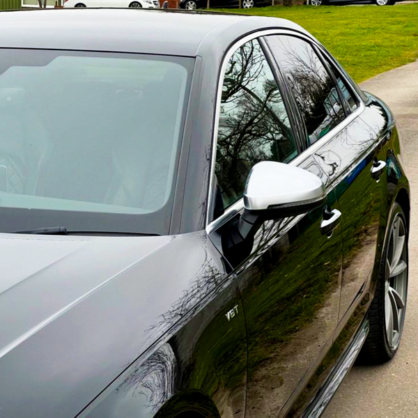 A sleek black car is parked on a paved road, reflecting trees on its polished surface, with "V6T" embossed near the front door. Grassy area with parked cars in the background.