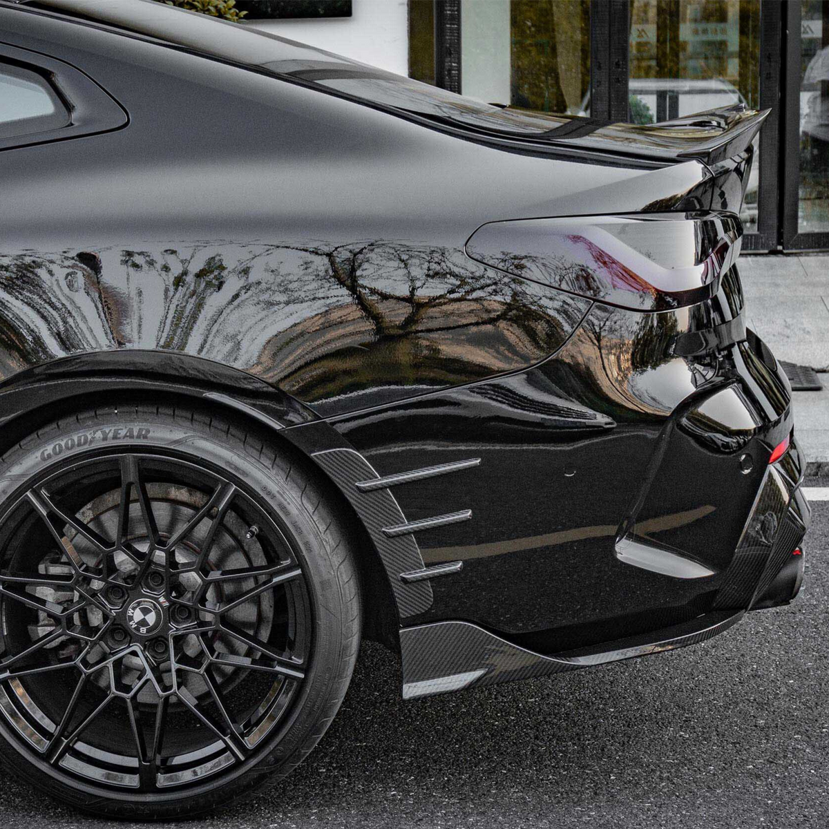 A sleek black sports car with intricate alloy wheels and sharp tail light design is parked outside a building. The tire displays the text "GOODYEAR."