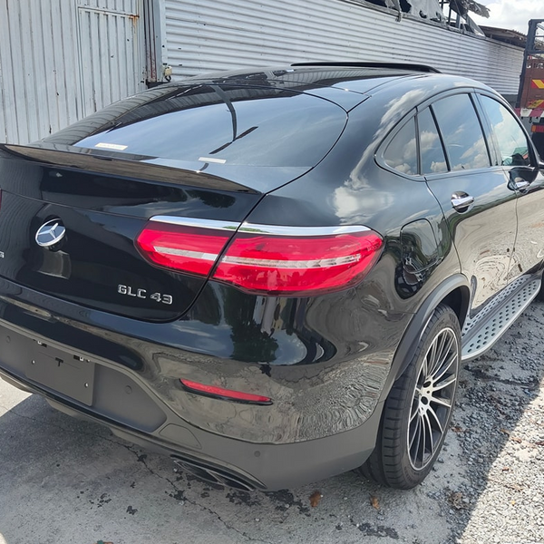 A black car labeled "GLC 43" and "AMG" is parked on gravel beside a corrugated metal wall and a truck. Bright sunlight casts shadows on the ground.