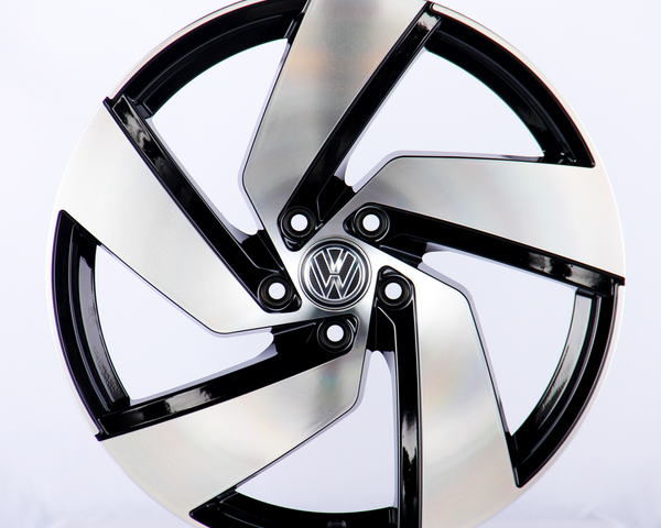 A sleek, silver and black alloy wheel with an intricate design sits upright in a well-lit studio. The central emblem features the Volkswagen logo against a plain white background.