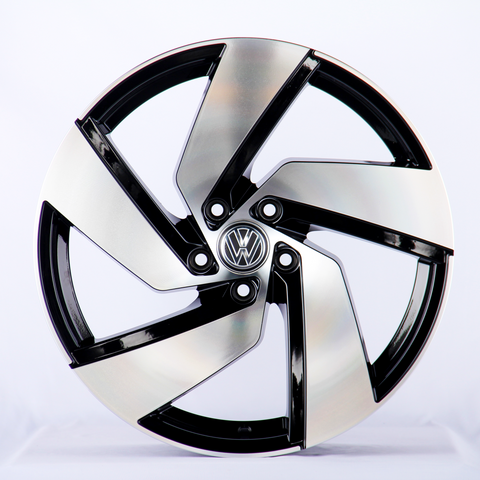 A sleek, silver and black alloy wheel with an intricate design sits upright in a well-lit studio. The central emblem features the Volkswagen logo against a plain white background.
