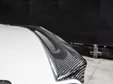 A carbon fiber car spoiler is attached to a white car, extending slightly upward. It is set against a dark, blurred background in an indoor setting.