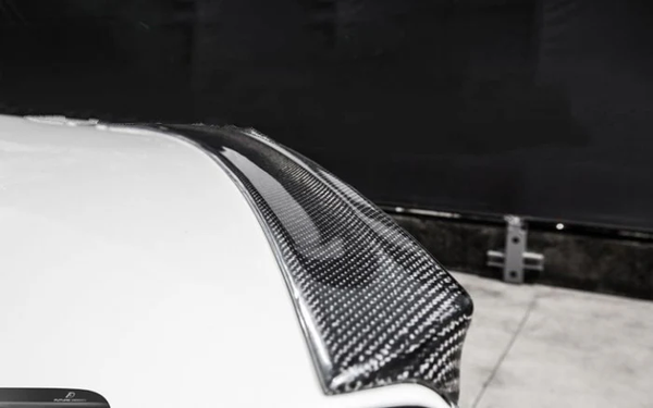 A carbon fiber car spoiler is attached to a white car, extending slightly upward. It is set against a dark, blurred background in an indoor setting.