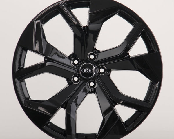 A glossy black car wheel rim with five split spokes, featuring the Audi logo in the center, is displayed against a plain white background.