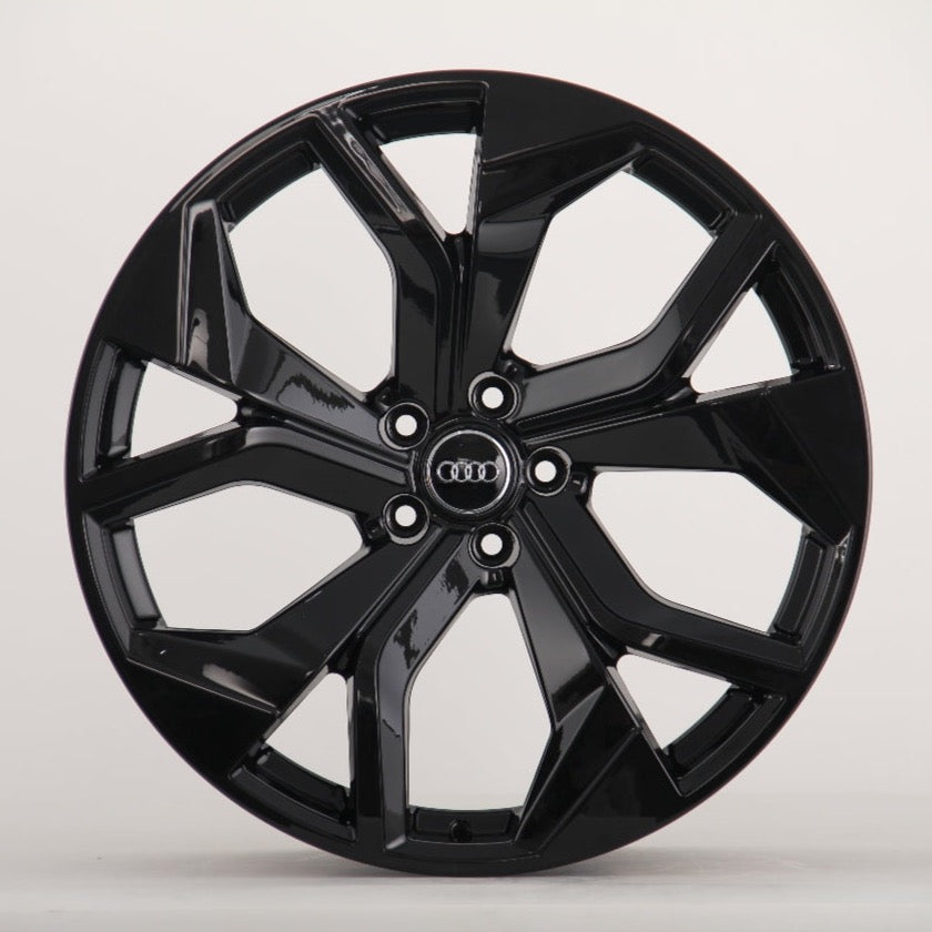 A glossy black car wheel rim with five split spokes, featuring the Audi logo in the center, is displayed against a plain white background.