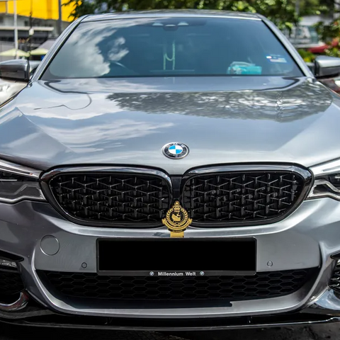 A silver BMW car is parked outdoors, displaying a gold emblem on the grille. Reflections of clouds are visible on its hood, with trees and buildings in the background. Number plate reads "Millennium Welt."