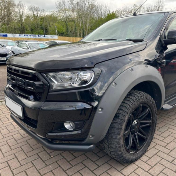 A black rugged pickup truck is parked on a paved lot, featuring chunky tires and a modified front grille. Other cars and trees are visible in the background.